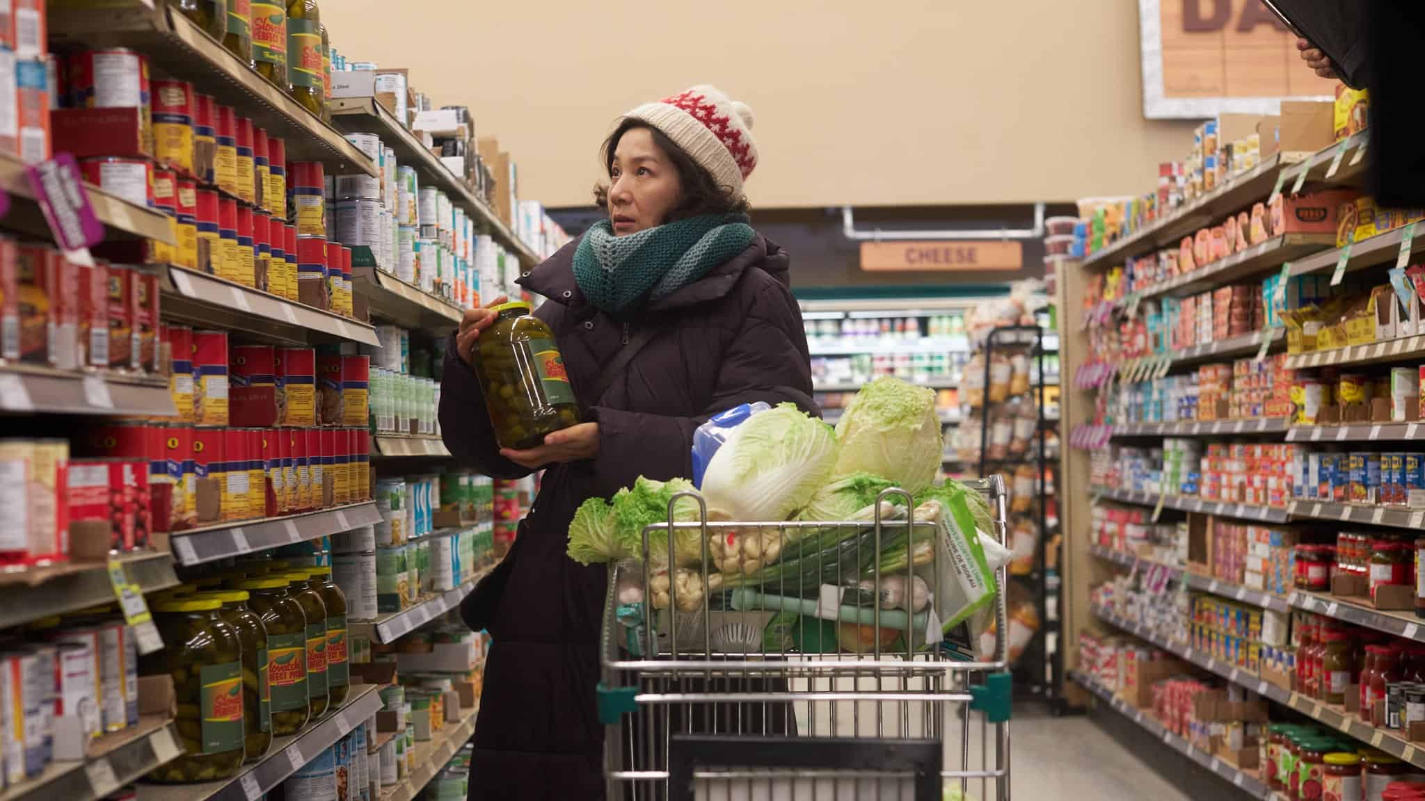 A person wearing a winter coat, scarf, and hat stands in a grocery store aisle, holding a large jar and looking at shelves. Their shopping cart is full of groceries, including leafy greens and cabbage.