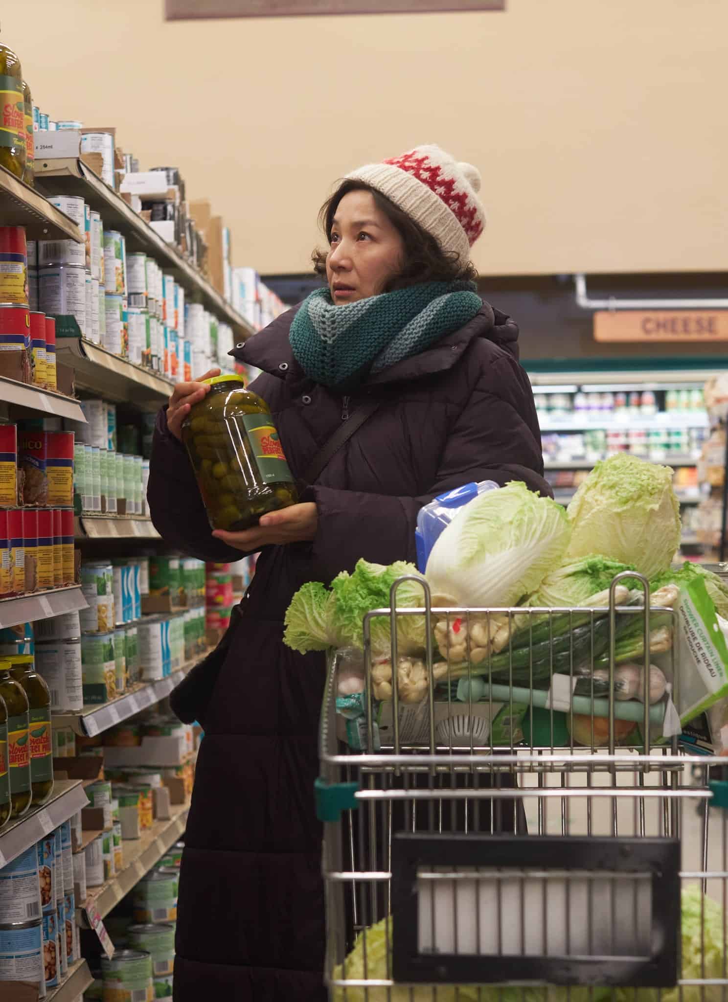 A person wearing a winter coat, scarf, and hat stands in a grocery store aisle, holding a large jar and looking at shelves. Their shopping cart is full of groceries, including leafy greens and cabbage.