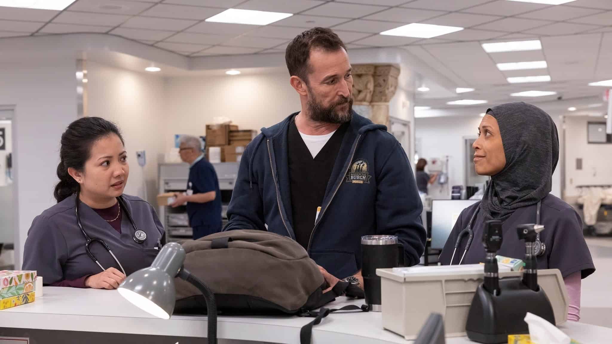 Three healthcare professionals stand and talk at a hospital reception desk. One woman wears a hijab and another has a stethoscope; a man in a hoodie stands between them. The setting is bright and clinical.
