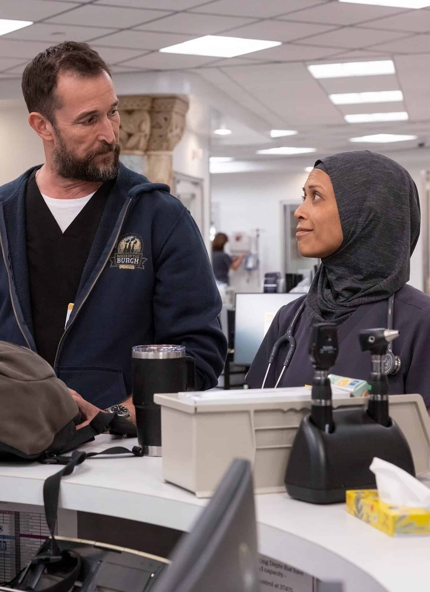 Three healthcare professionals stand and talk at a hospital reception desk. One woman wears a hijab and another has a stethoscope; a man in a hoodie stands between them. The setting is bright and clinical.
