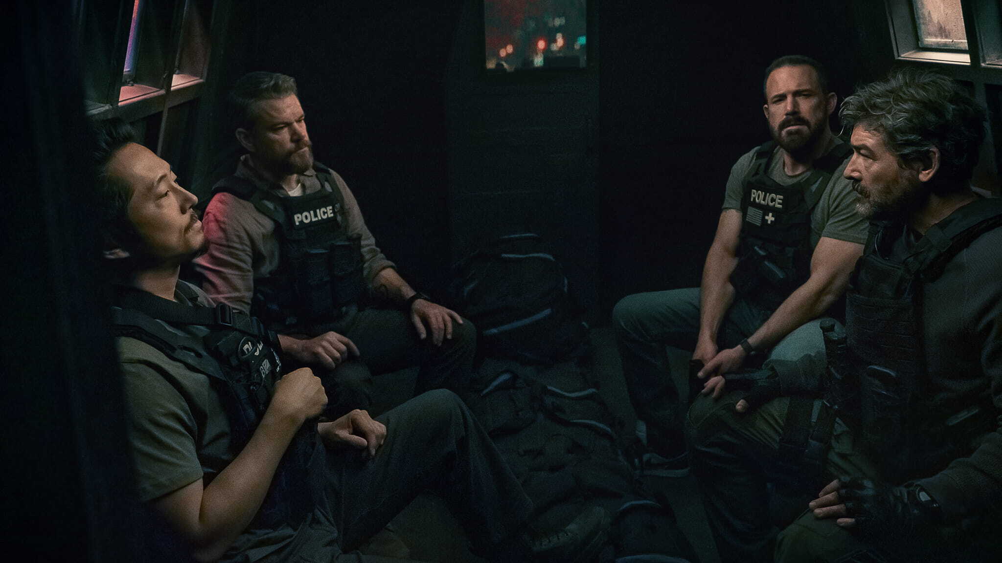Four men in tactical police gear sit inside a dimly lit armored vehicle, appearing tense and deep in thought, with gear bags on the floor between them.