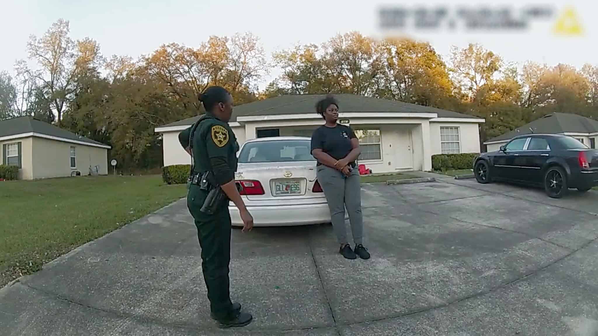 A police officer stands beside a woman who is leaning against the trunk of a white car parked in a residential driveway. A house and another parked car are visible in the background.
