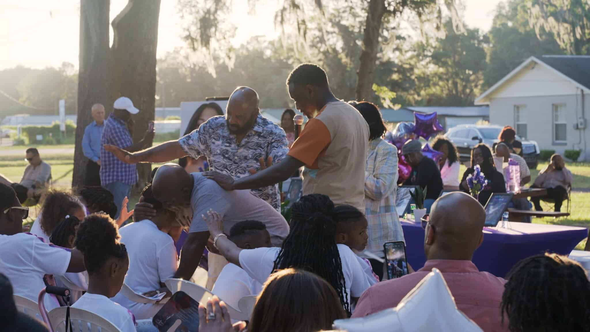 A lively outdoor gathering where a man dances energetically among seated people, while others watch, smile, and interact. Sunlight filters through trees and balloons decorate the background.