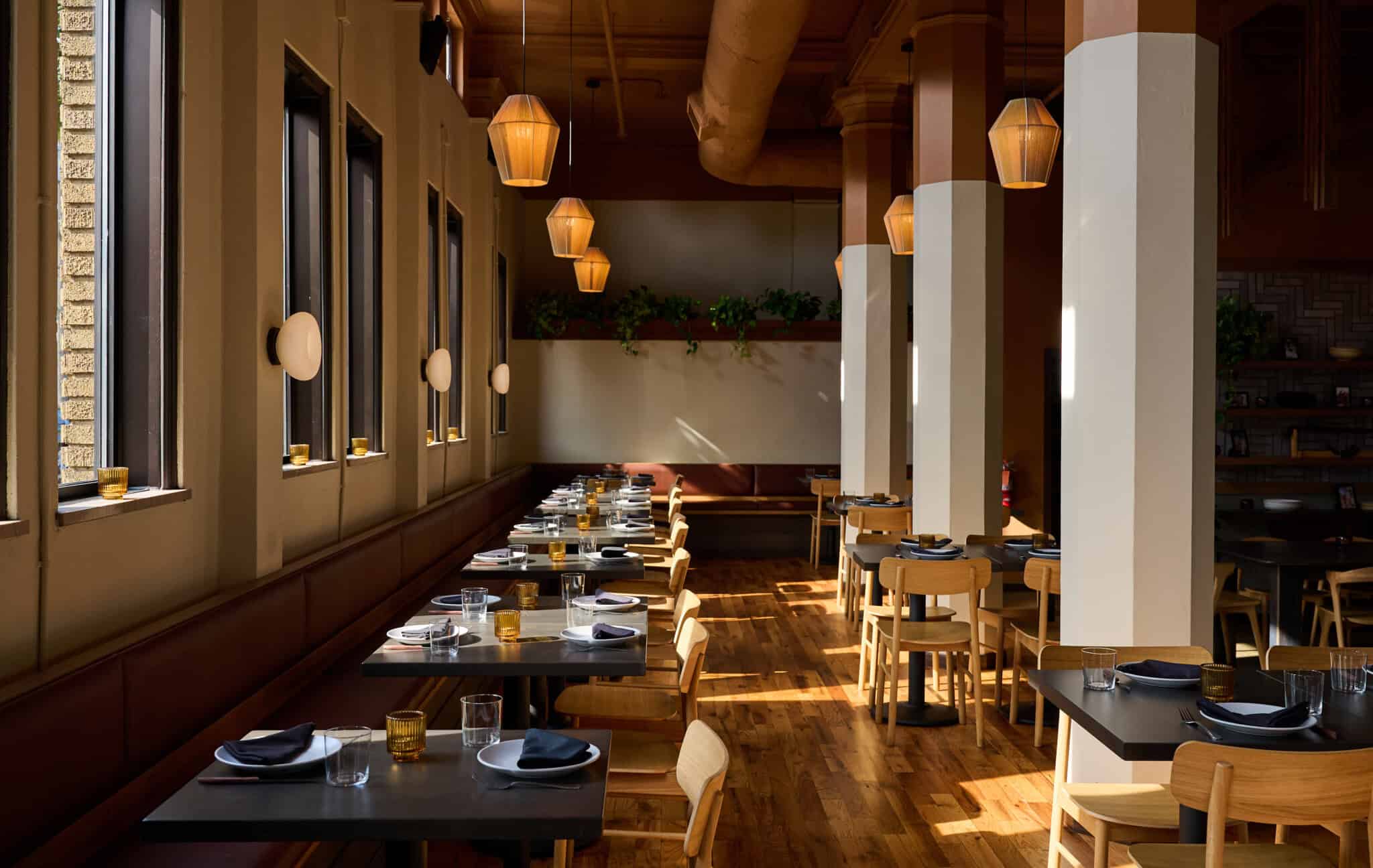 A modern restaurant with wooden floors, neatly set tables, wooden chairs, large windows letting in natural light, hanging pendant lights, and green plants on a wall shelf in the background.
