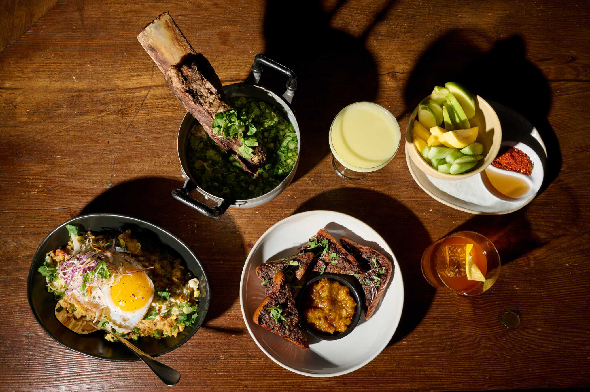 A wooden table with various dishes: a bowl with rice and a sunny-side-up egg, a pot with ribs and green garnish, a plate of meat and vegetables, a bowl of sliced fruit, and two drinks, one yellow and one amber.