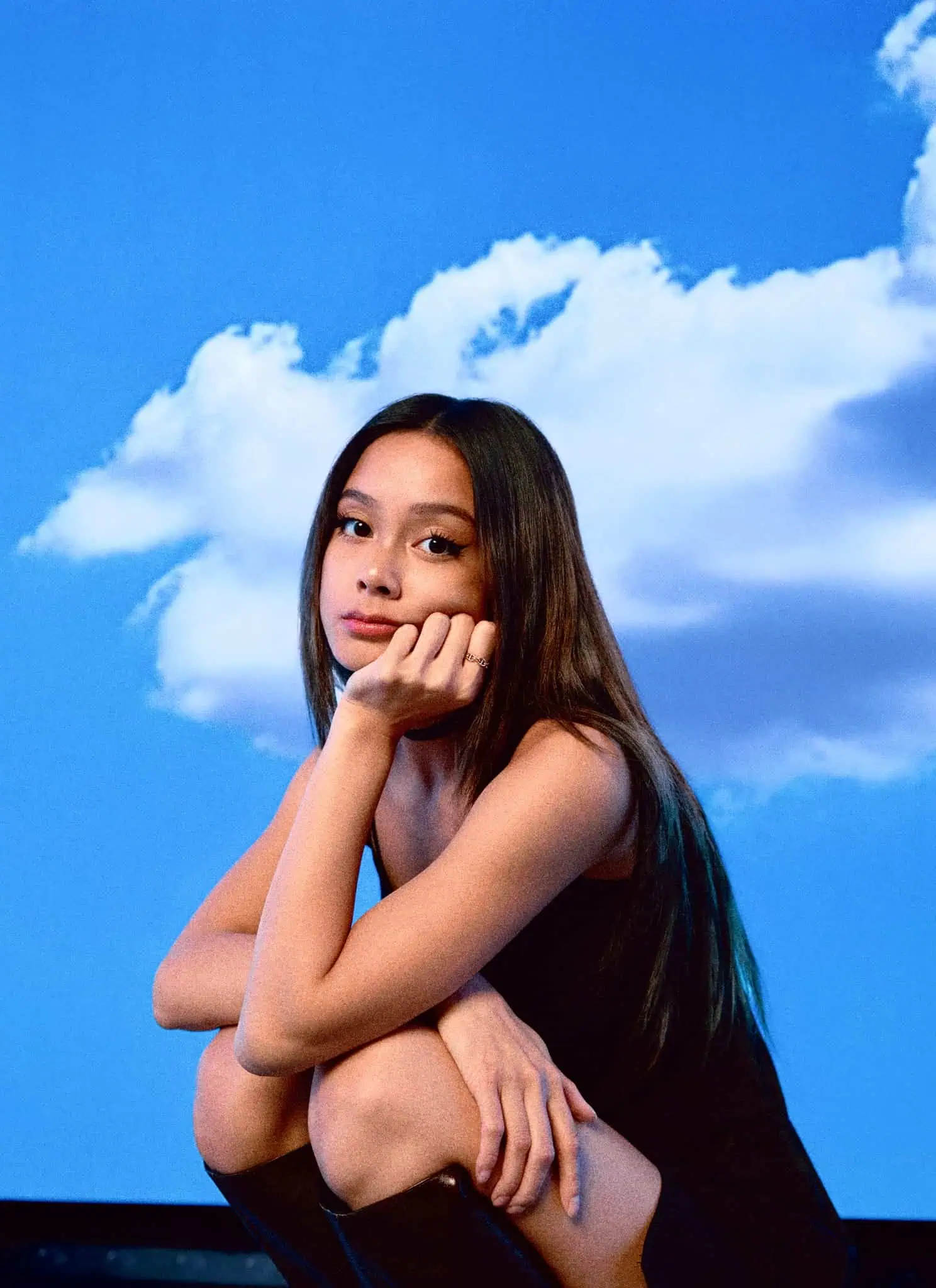 A person with long dark hair in a black dress and boots squats with their chin resting on one hand, set against a bright blue sky background with scattered white clouds.