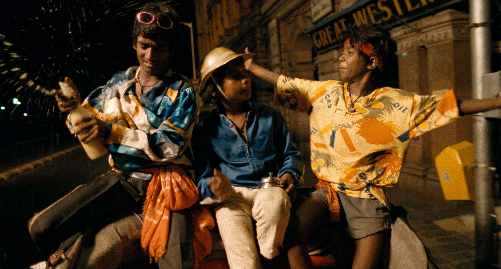 Three boys in colorful clothes sit together on a cart at night—one sprays a bottle, another wears a gold helmet, and the third stretches out his arms. Their lively scene on a city street feels like a moment from a Mira Nair film.