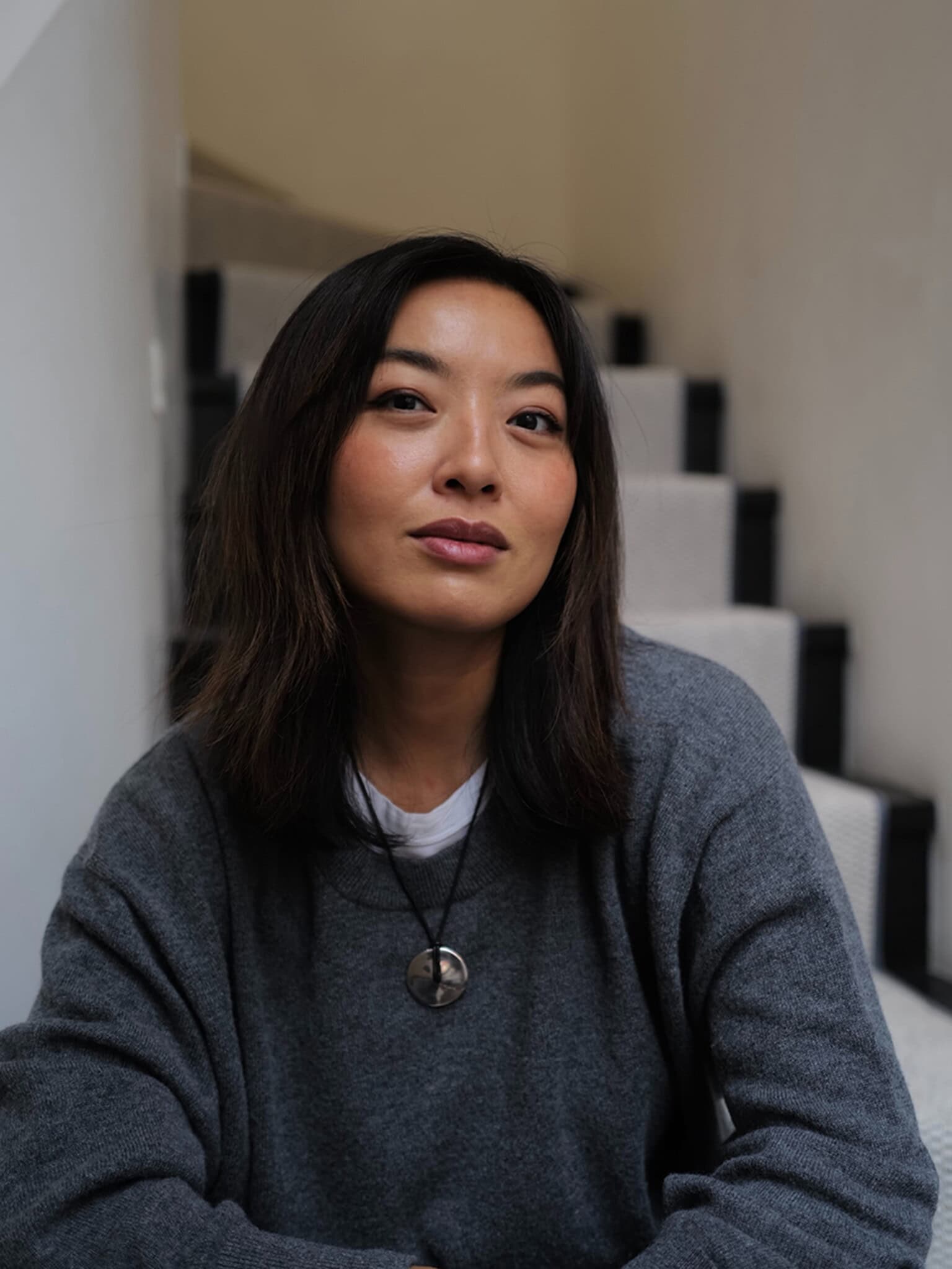 A woman with shoulder-length dark hair, wearing a grey sweater and a round pendant necklace, sits on indoor stairs looking confidently at the camera. The background is softly lit and neutral.