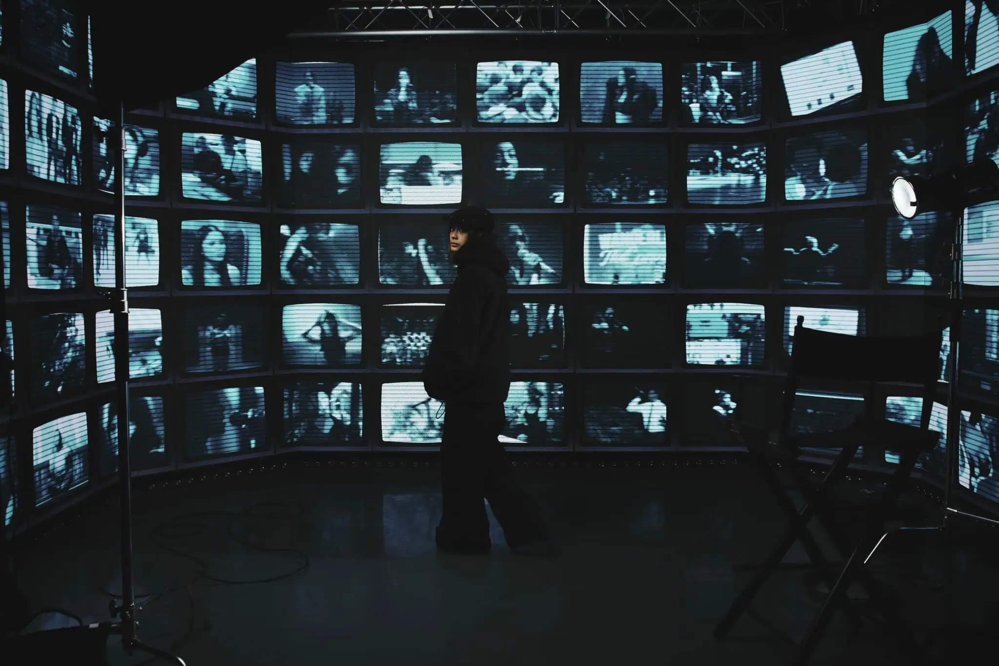 A person stands in front of a curved wall of TV screens displaying various black-and-white images, creating a surveillance or control room atmosphere. A director’s chair and studio lights are visible.