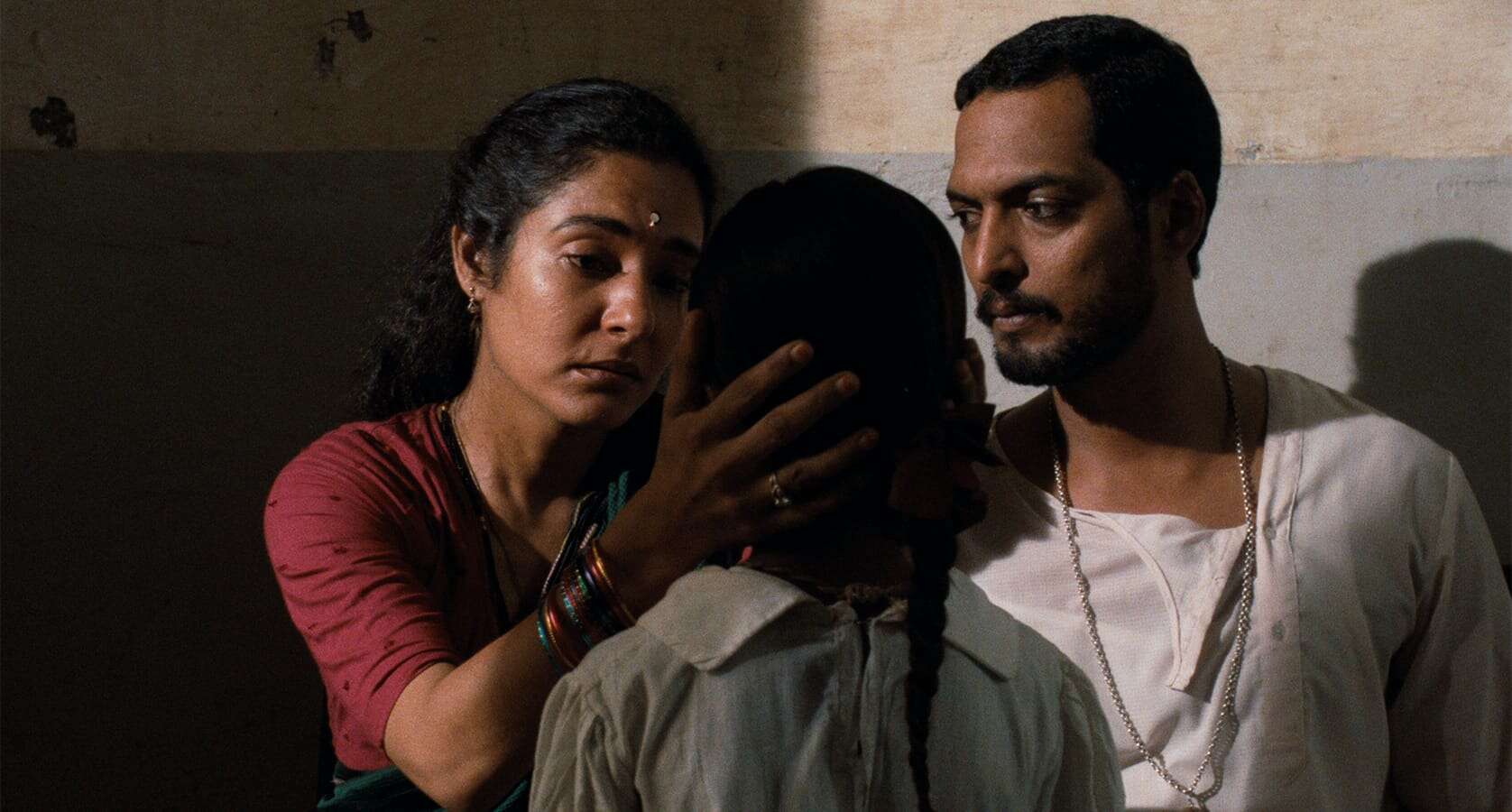 A woman gently holds a girl’s face, looking at her with concern, while a man stands beside them watching—a scene reminiscent of Mira Nair’s evocative storytelling. The setting appears to be indoors with plain walls in the background.