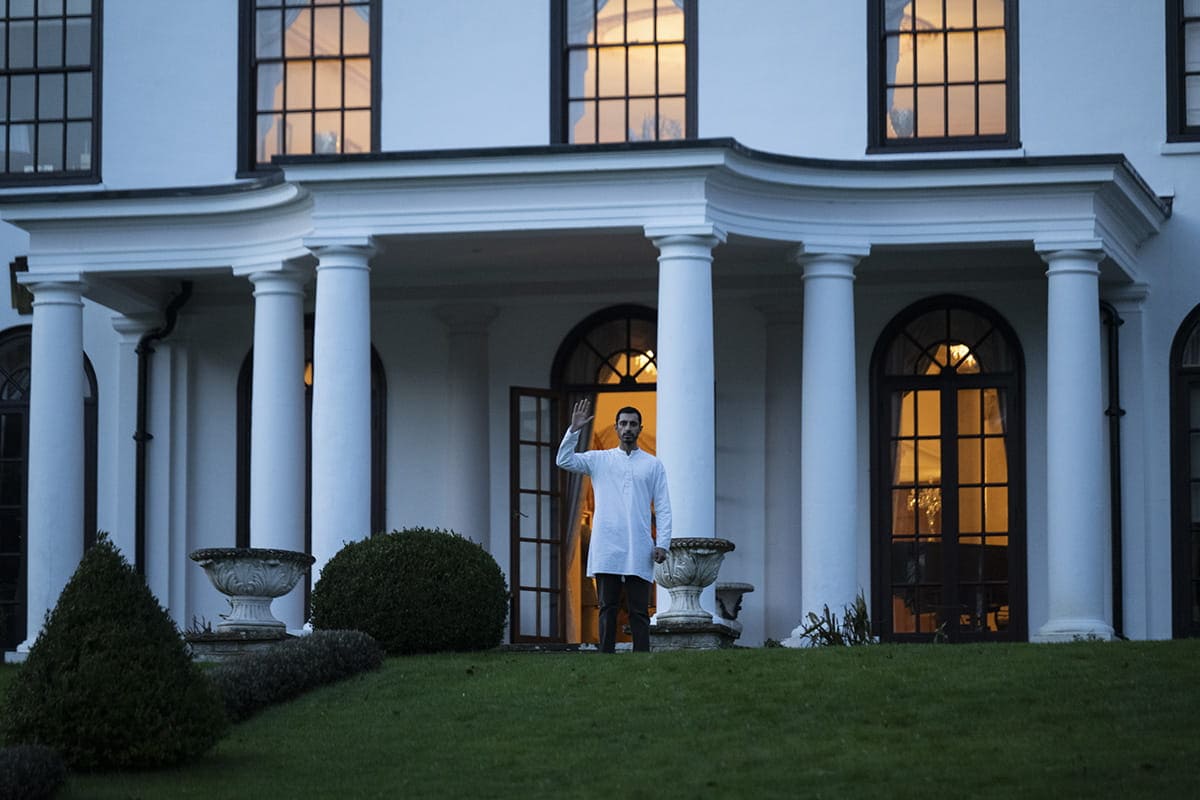 A man in a white shirt stands in front of a large white building with tall columns and arched windows, waving toward the camera as evening light glows from inside.