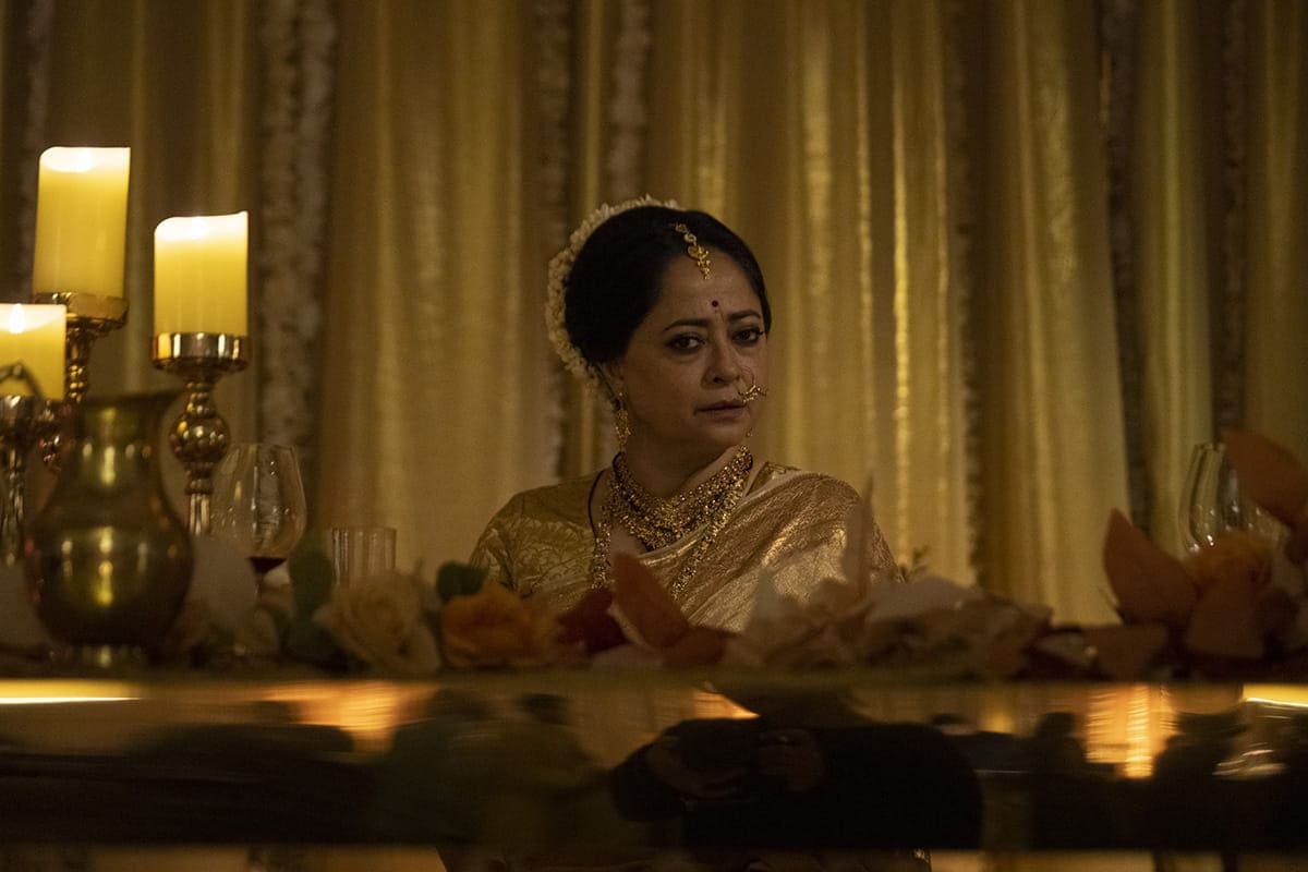 A woman in traditional Indian attire and jewelry sits at a decorated table with candles, flowers, and glassware, against a gold curtain backdrop.
