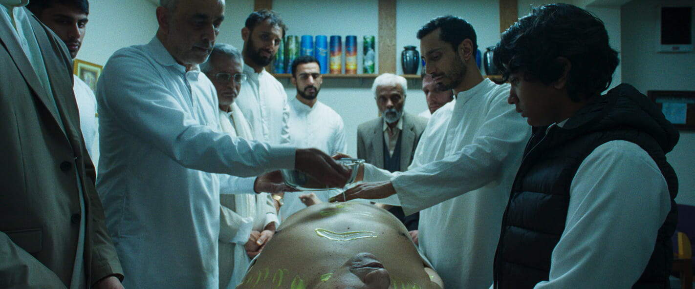 A group of men, some in white coats and some in regular clothes, stand around a body on a table in a brightly lit room. One man pours water on the body, while others observe closely, appearing solemn and serious.