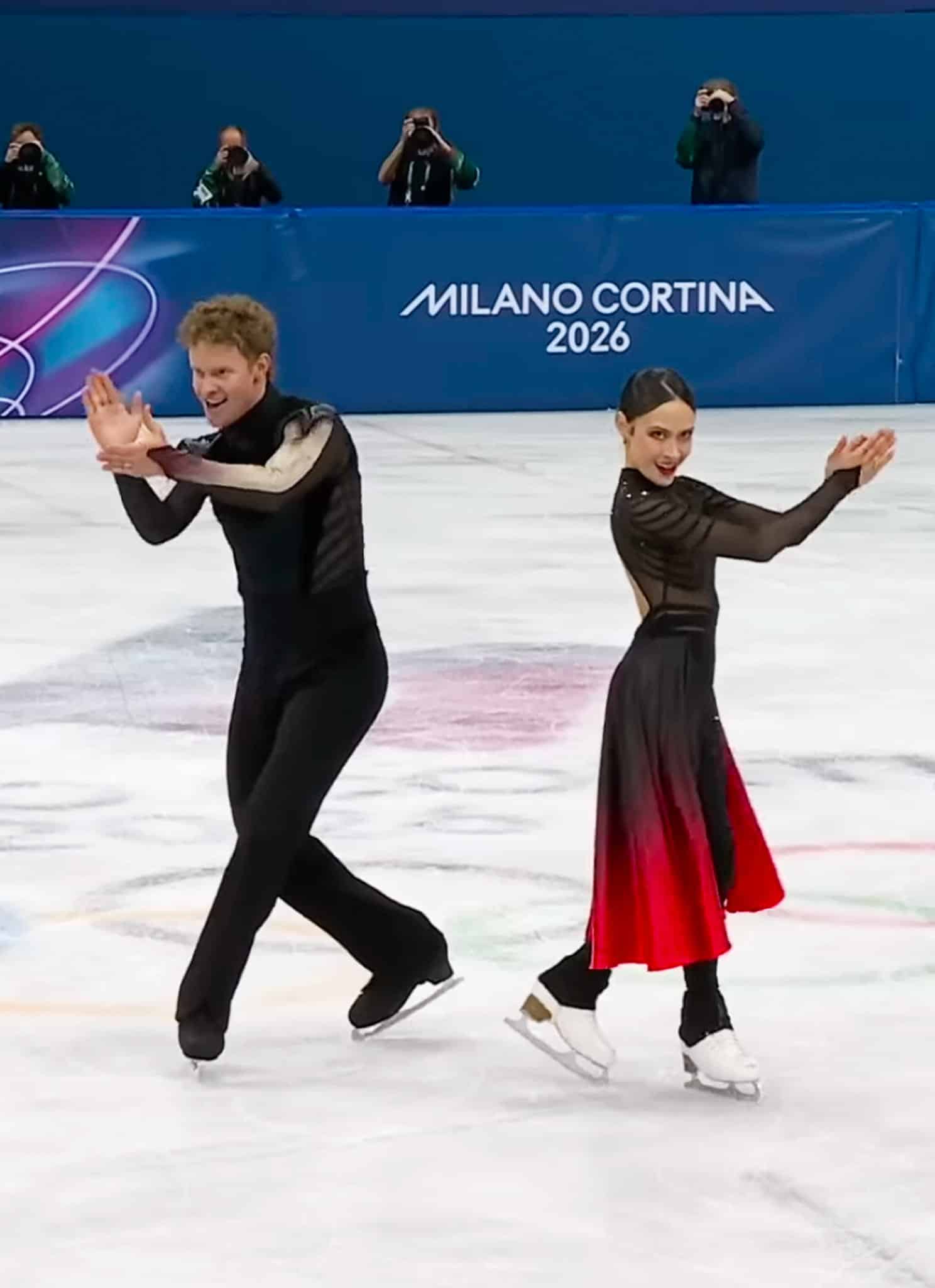 Two figure skaters perform a synchronized routine on ice, facing opposite directions with hands raised. A Milano Cortina 2026 banner is visible behind them, with photographers capturing the moment.