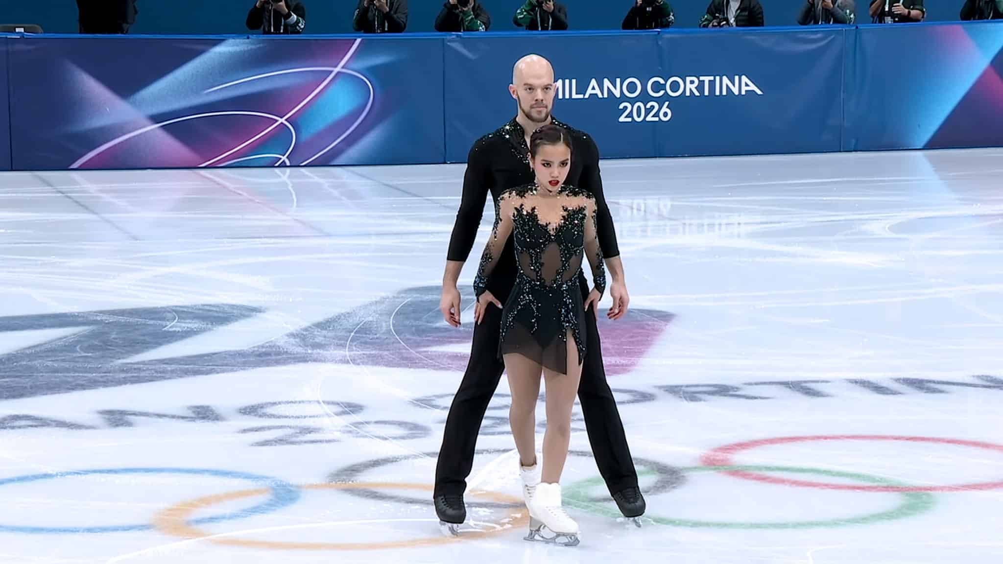 Two figure skaters, a man and a woman, perform on ice in dramatic black costumes. The ice rink displays Milano Cortina 2026 and Olympic rings in the background, with photographers visible along the boards.