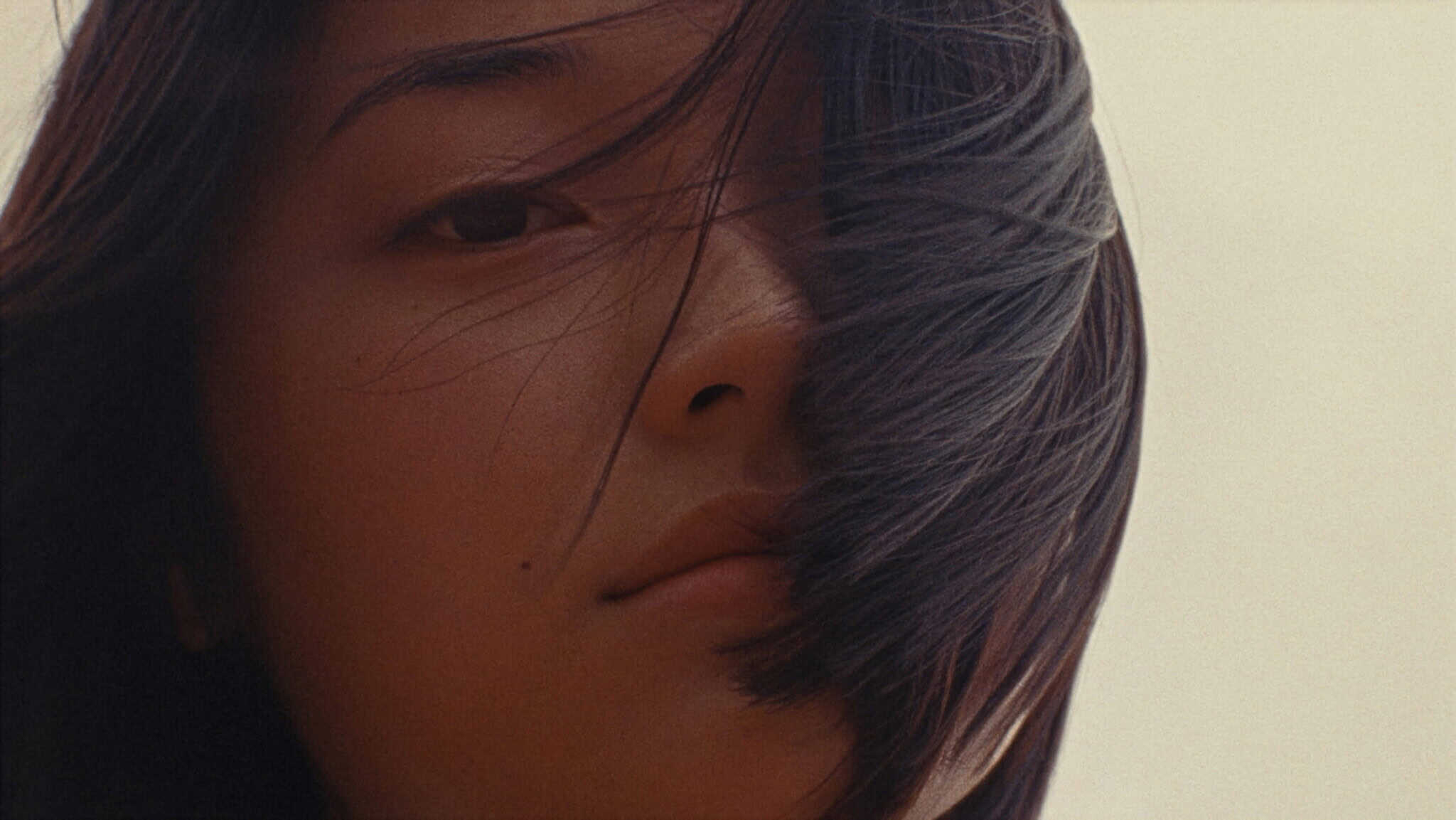 Close-up of a woman’s face with straight dark hair partially covering one eye. Her expression is neutral and she is looking into the camera. The background is softly blurred and light in color.