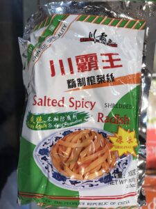 A package of Salted Spicy Shredded Radish from China, reminiscent of Asian childhood snacks, featuring an image of shredded radish on a plate. The packaging is predominantly green, white, and red with both English and Chinese text.
