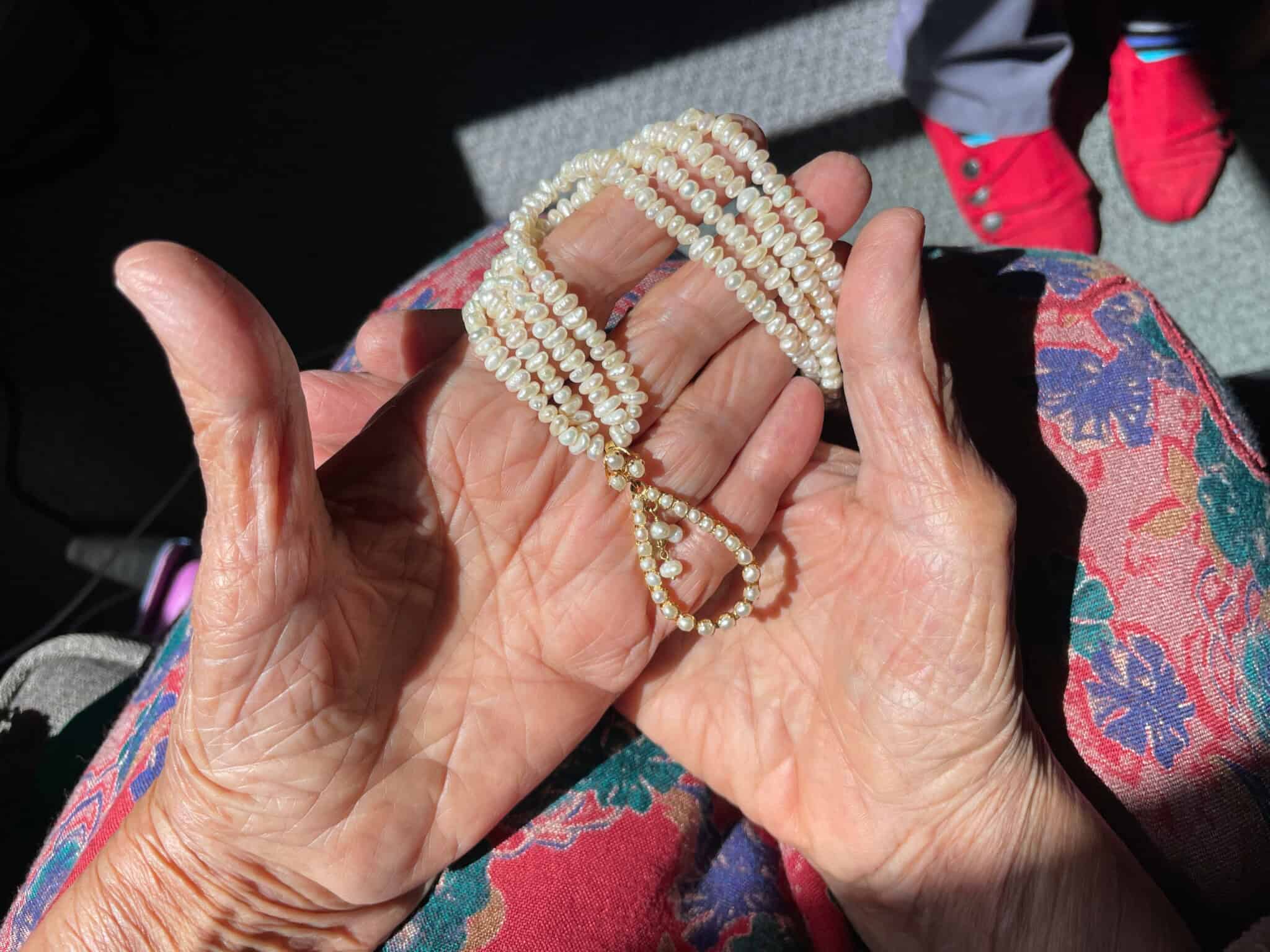 A pair of elderly hands holds a multi-strand pearl necklace. The person wears a colorful floral-patterned garment, and red shoes are visible in the background under bright sunlight.