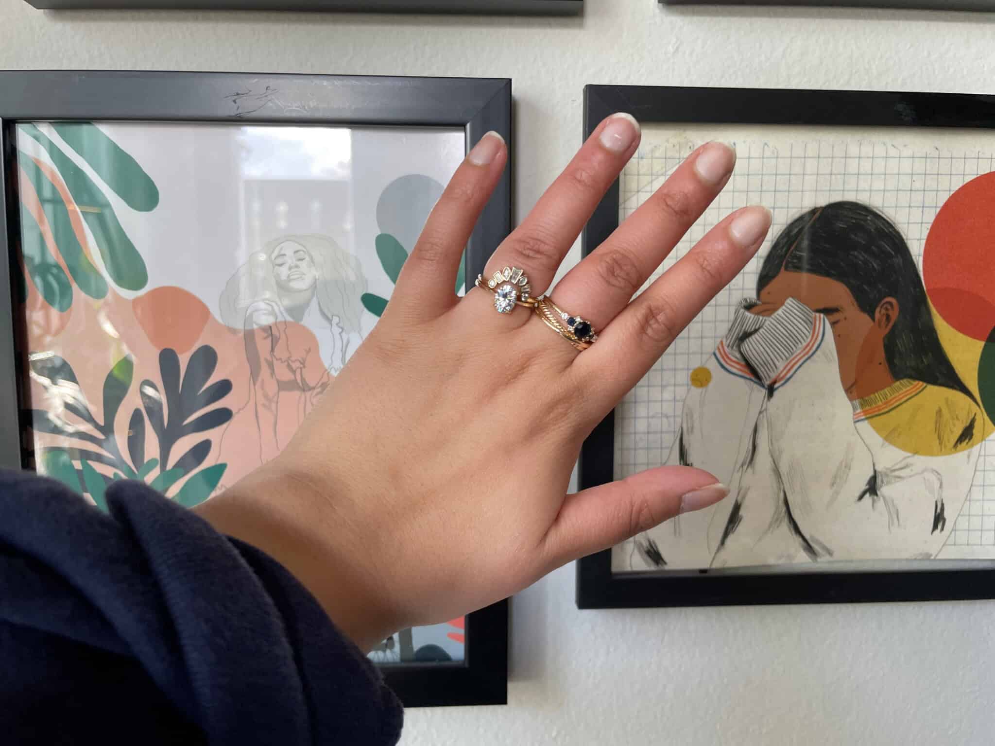 A hand with manicured nails and three rings is held up in front of two framed art pieces on a white wall. One artwork shows a person singing, and the other shows a person covering their face.