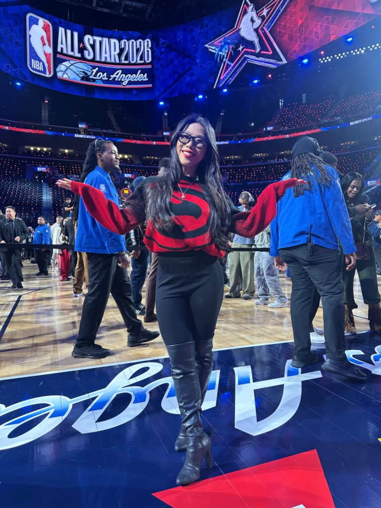 A woman in a red and black sweater, black pants, and knee-high boots stands smiling on a basketball court, arms outstretched. NBA All-Star 2026 signs and people are visible in the background.