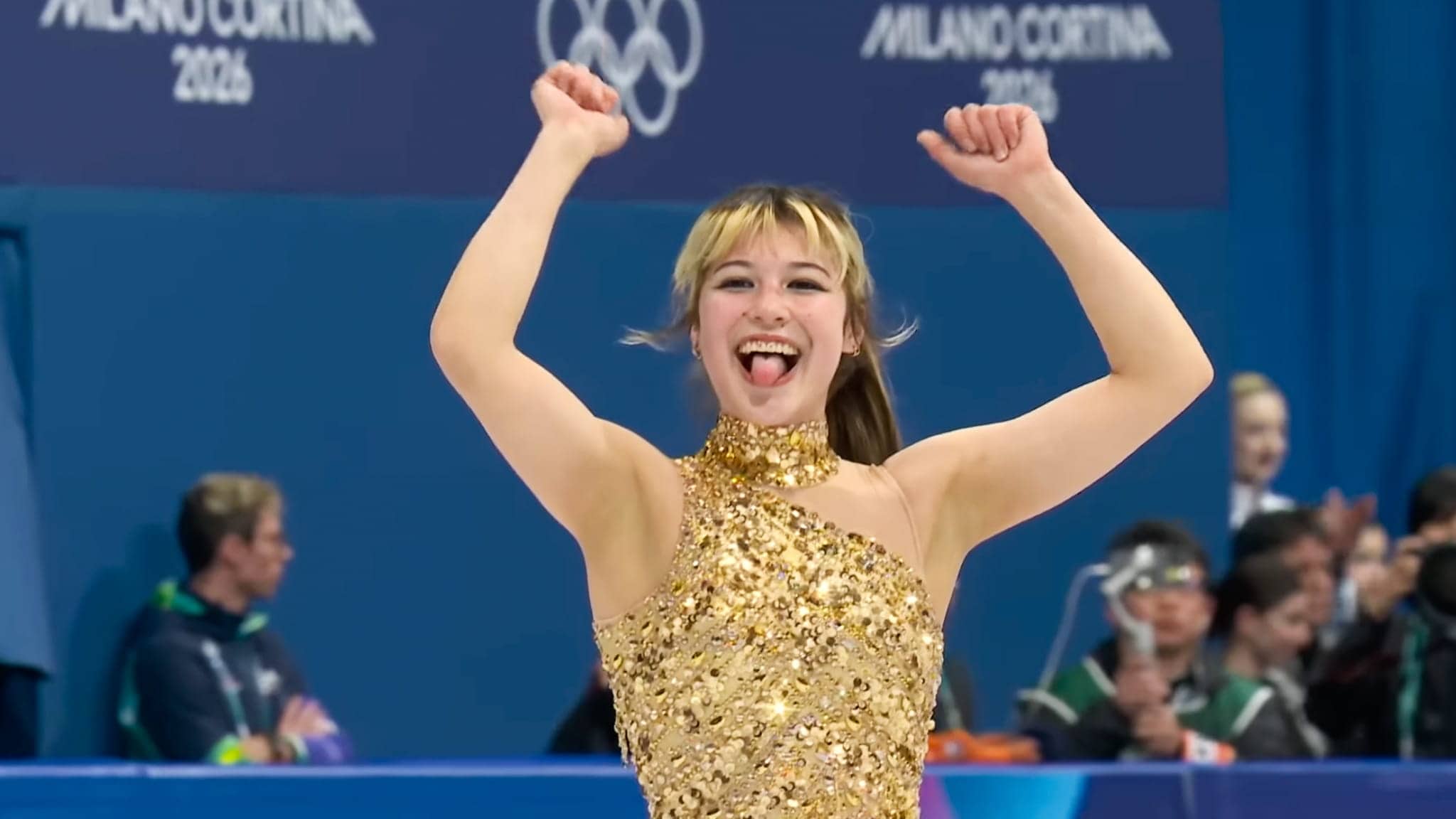 A figure skater in a sparkling gold costume smiles with arms raised and tongue out in celebration, with Olympic rings and Milano Cortina 2026 in the background. Spectators are visible behind her.