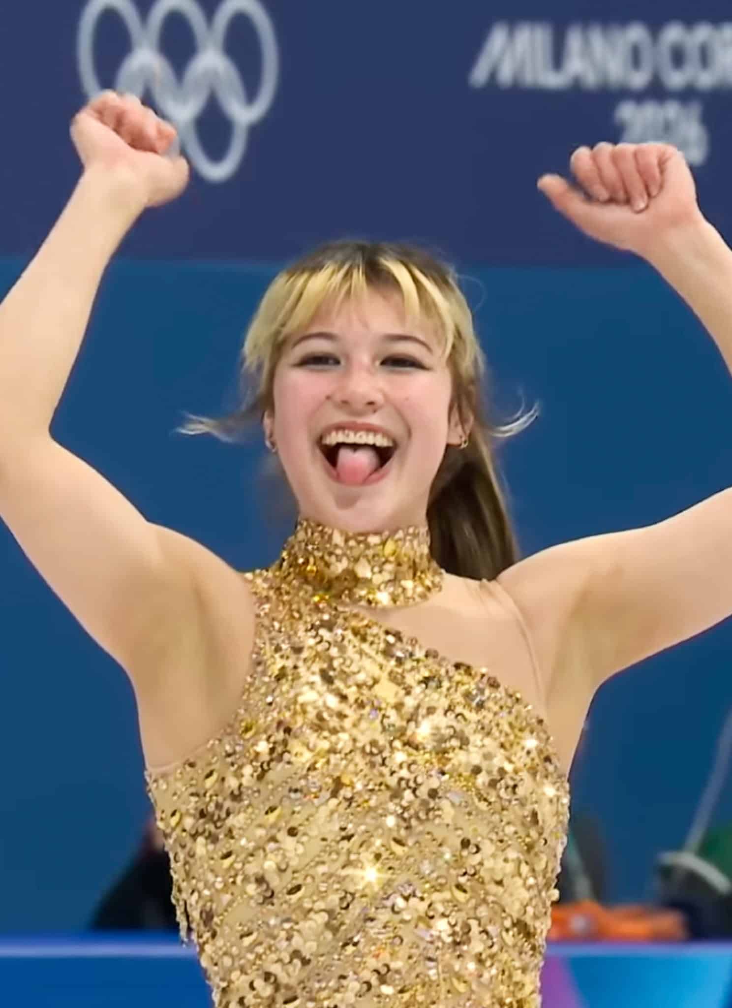 A figure skater in a sparkling gold costume smiles with arms raised and tongue out in celebration, with Olympic rings and Milano Cortina 2026 in the background. Spectators are visible behind her.