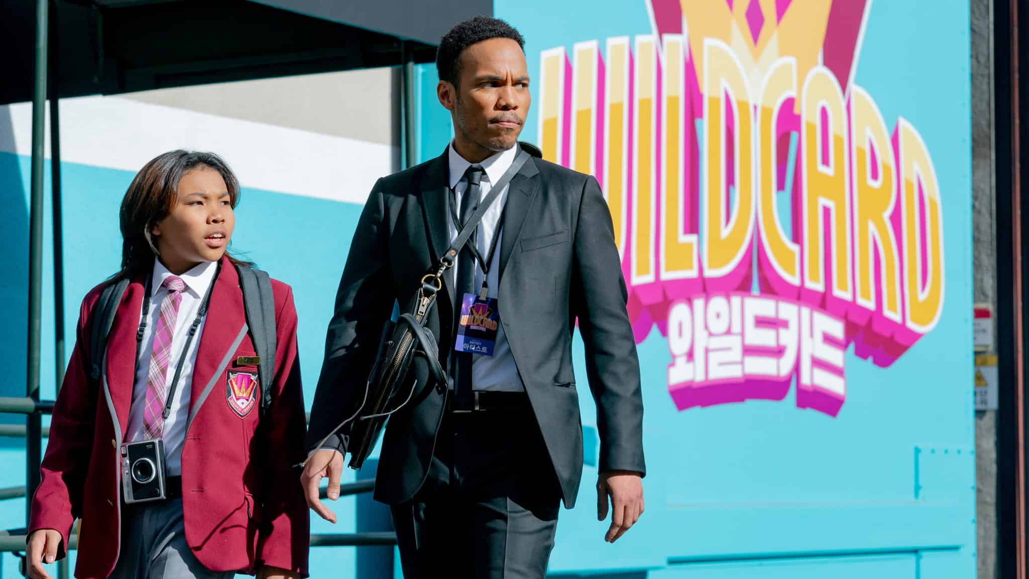 A man in a suit and a child in a red school uniform walk outside past a colorful wall with the word WILDCARD in bold letters and Korean text below it. Both have serious expressions and are carrying bags.