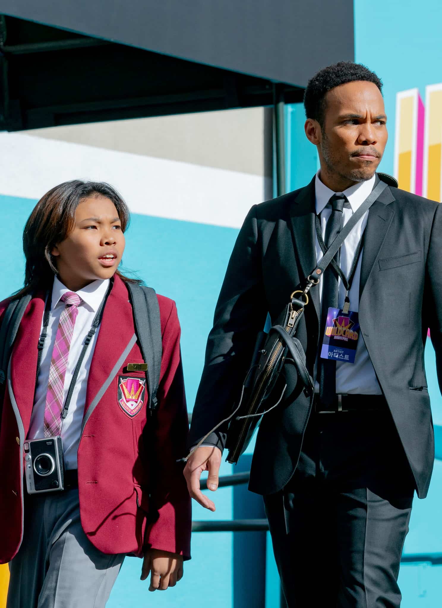 A man in a suit and a child in a red school uniform walk outside past a colorful wall with the word WILDCARD in bold letters and Korean text below it. Both have serious expressions and are carrying bags.
