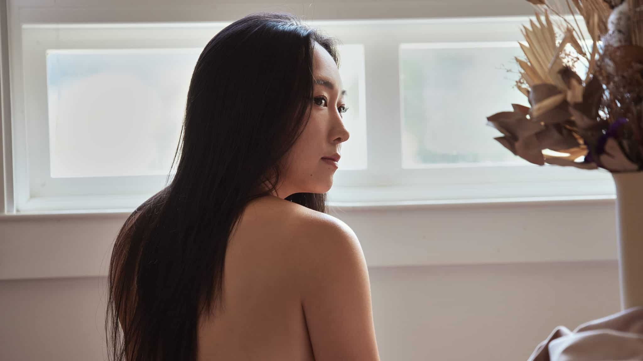 A woman with long dark hair sits with her back exposed, looking over her shoulder toward a window. Natural light illuminates her face, and dried flowers in a vase are visible beside her.