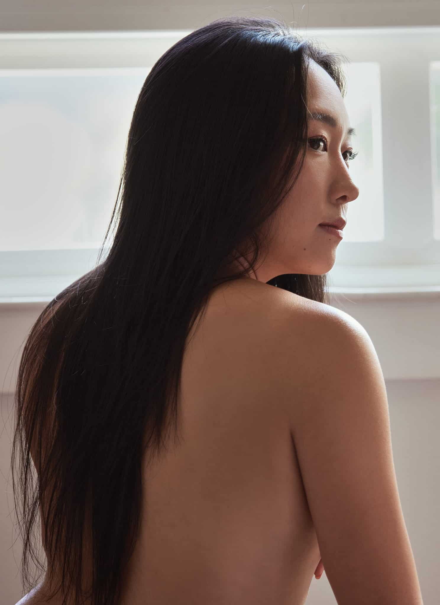 A woman with long dark hair sits with her back exposed, looking over her shoulder toward a window. Natural light illuminates her face, and dried flowers in a vase are visible beside her.