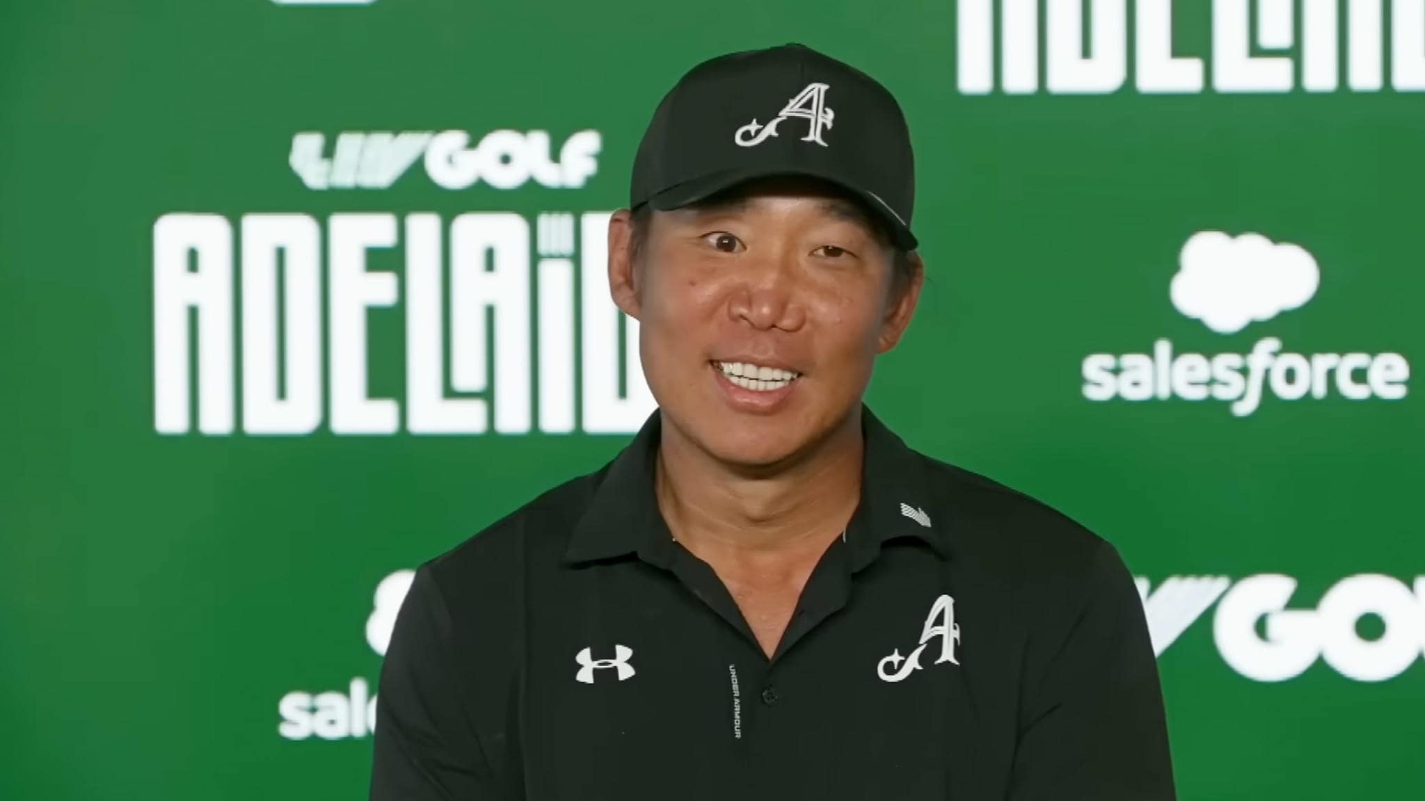 A man wearing a black cap and shirt with an A logo sits in front of a green backdrop featuring LIV Golf Adelaide and Salesforce logos, smiling at the camera.