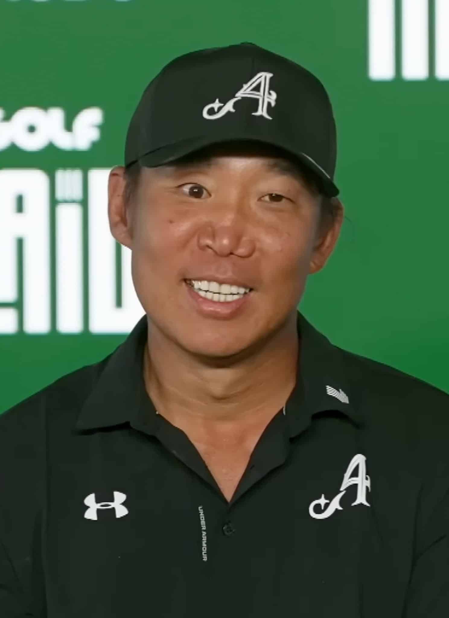 A man wearing a black cap and shirt with an A logo sits in front of a green backdrop featuring LIV Golf Adelaide and Salesforce logos, smiling at the camera.