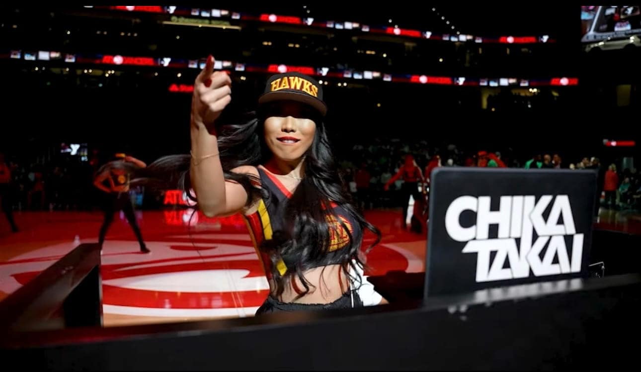 A DJ wearing an Atlanta Hawks cap and jersey points towards the camera while performing at a basketball arena, with CHIKA TAKAI displayed on her DJ booth.