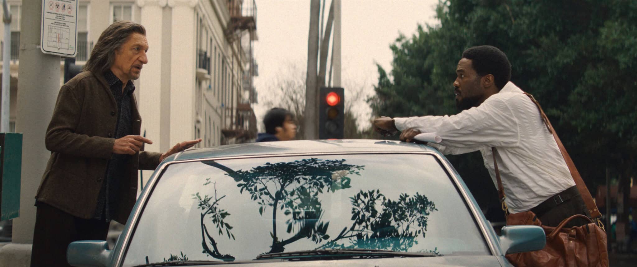 Two men stand on opposite sides of a car, talking intently on a city street. The car’s windshield has large artistic stickers of trees. A red traffic light and buildings are visible in the background.