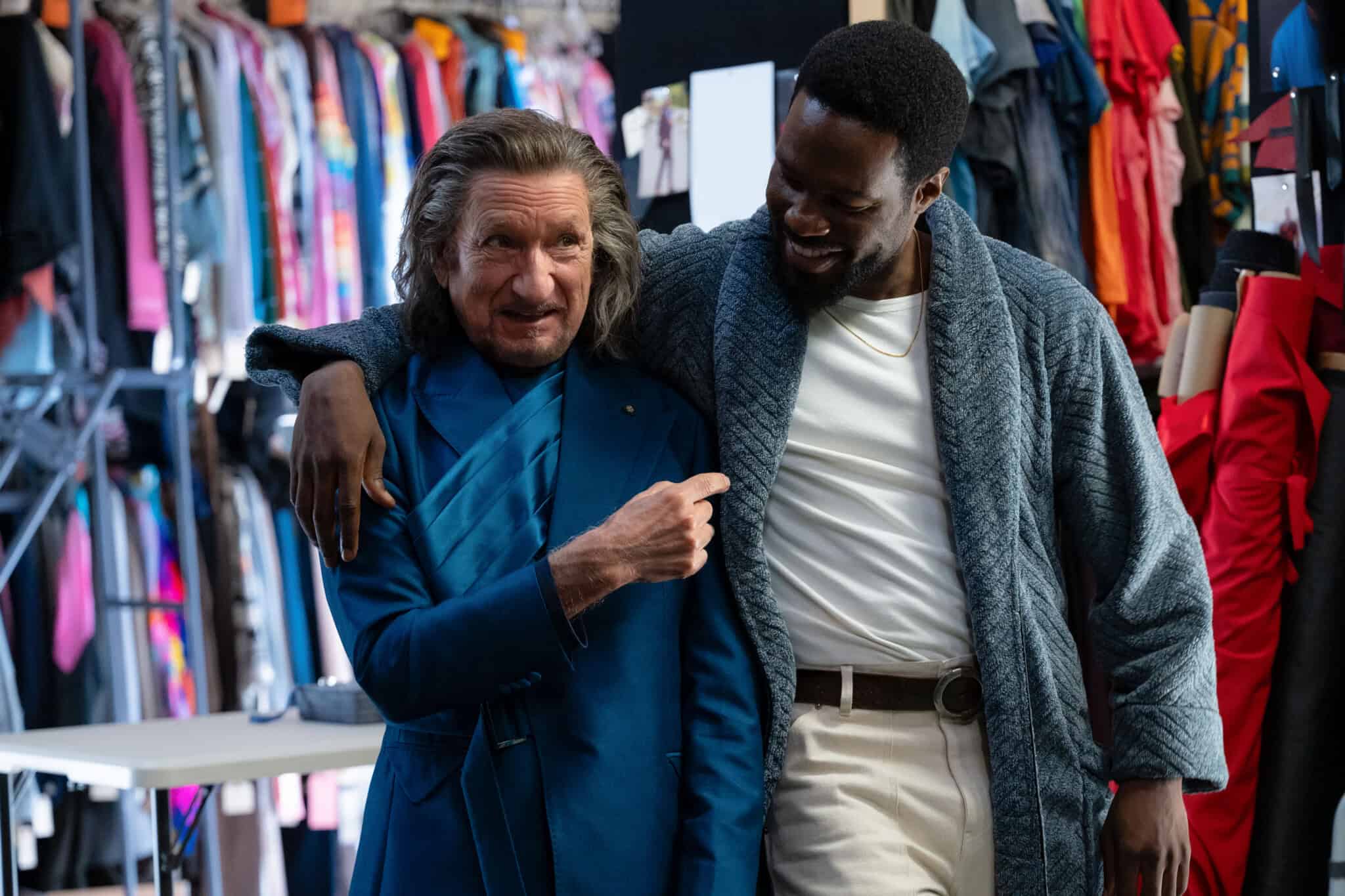 Two men stand together smiling in a colorful clothing store. The older man with long gray hair wears a blue blazer, while the younger man with short black hair and beard wears a blue robe and white shirt. They appear to be enjoying each other’s company.
