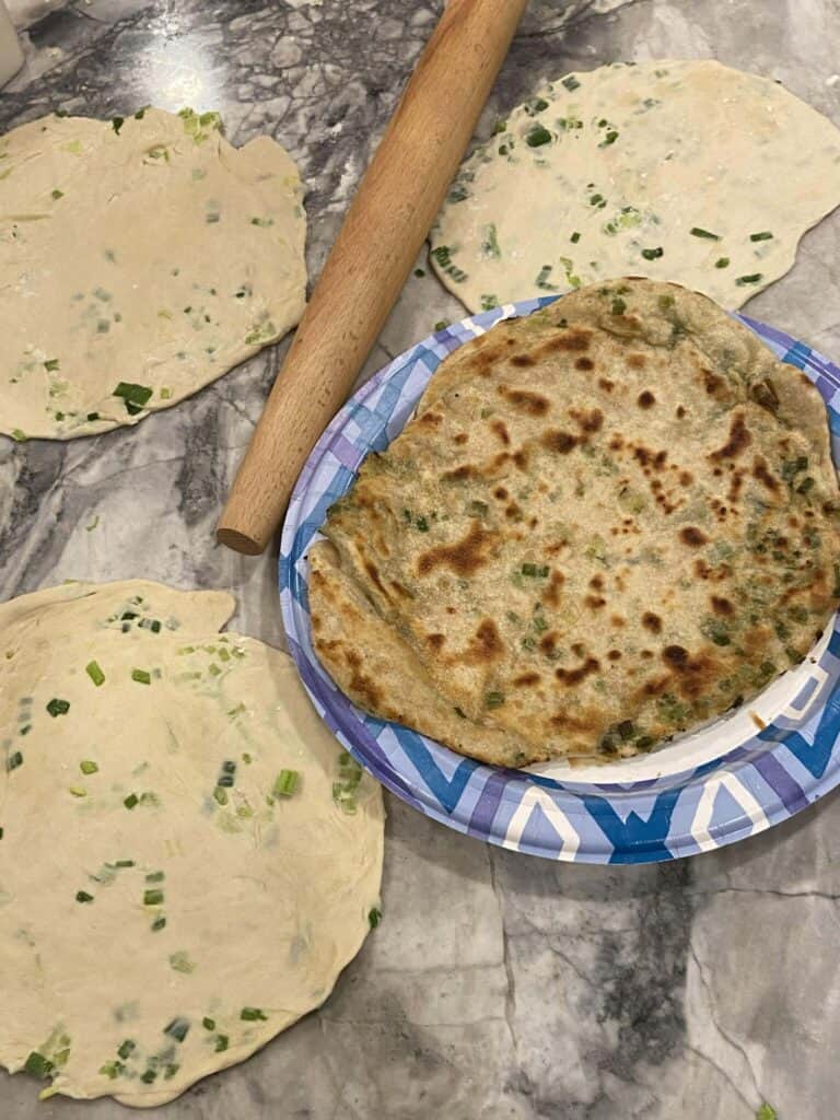 A marble countertop with rolled-out dough circles containing green onions, a rolling pin, and a cooked scallion pancake on a blue and white plate.