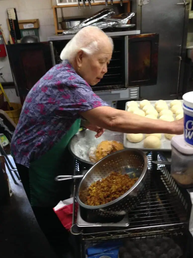 An elderly woman with white hair, wearing a floral shirt and green apron, prepares dough balls in a kitchen. In front of her are a colander with a cooked filling and a bucket labeled “CW.”.