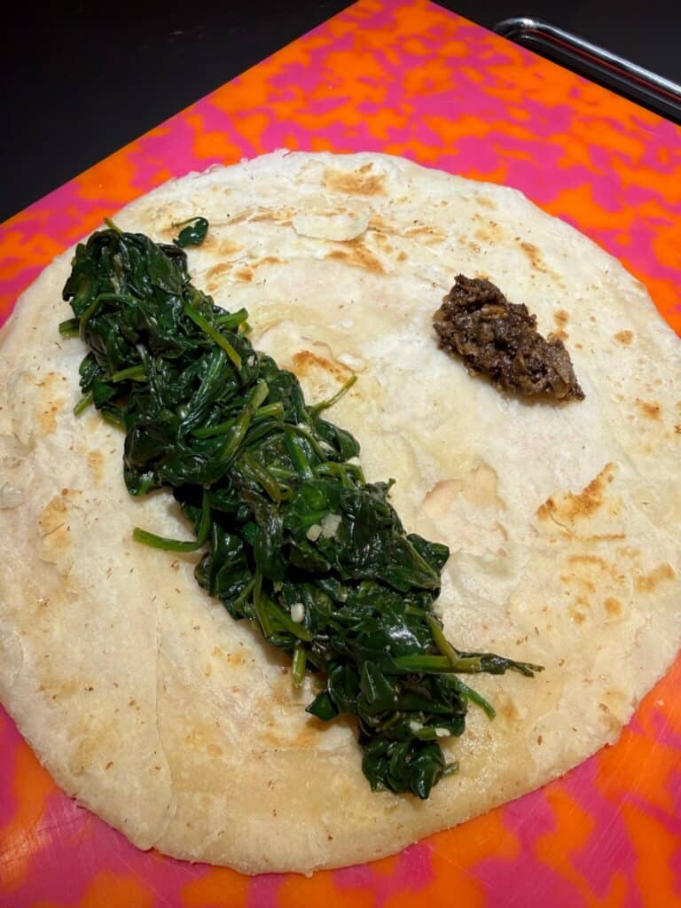 A large flatbread on a bright pink and orange cutting board, topped with a line of cooked spinach on one side and a small scoop of dark tapenade-like spread on the other side.