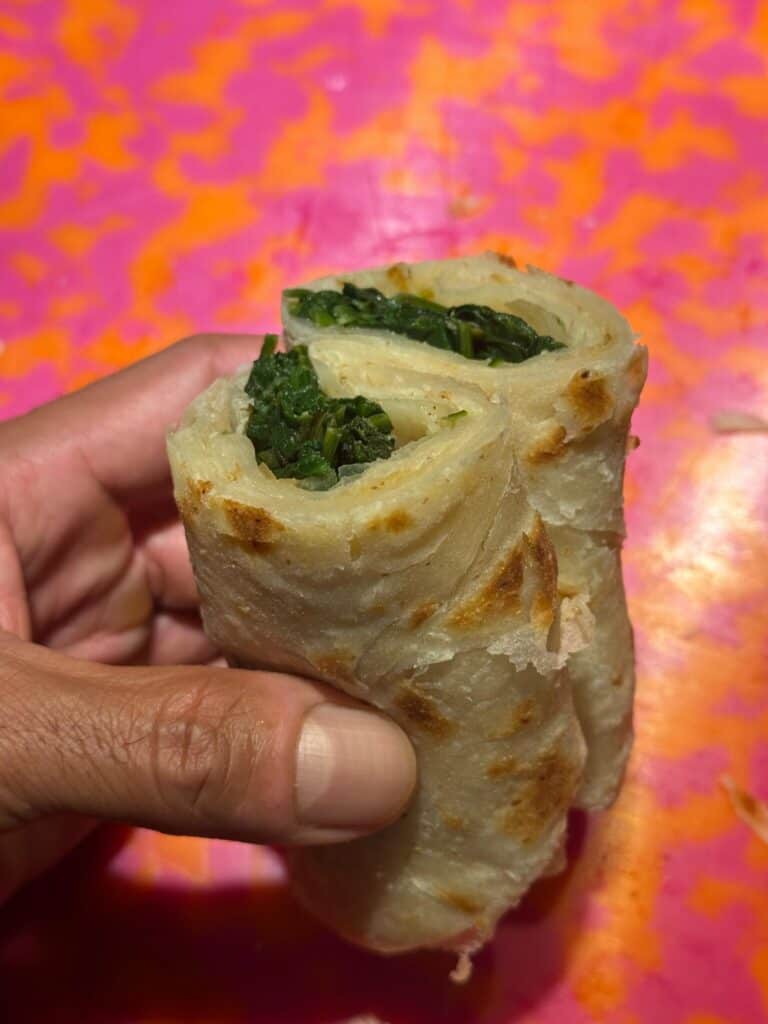 A hand holds a rolled flatbread filled with leafy green vegetables, against a bright pink and orange patterned background.