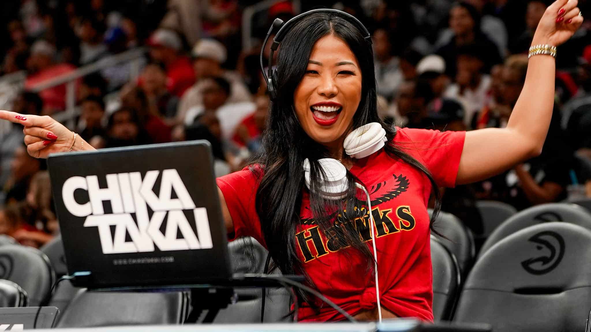 A smiling woman wearing a red Atlanta Hawks shirt and headphones poses energetically behind a DJ booth with a laptop labeled CHIKA TAKAI at a sports arena, with a crowd visible in the background.