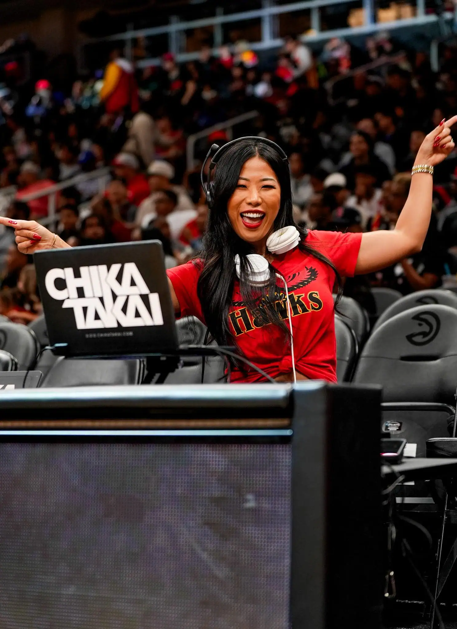 A smiling woman wearing a red Atlanta Hawks shirt and headphones poses energetically behind a DJ booth with a laptop labeled CHIKA TAKAI at a sports arena, with a crowd visible in the background.