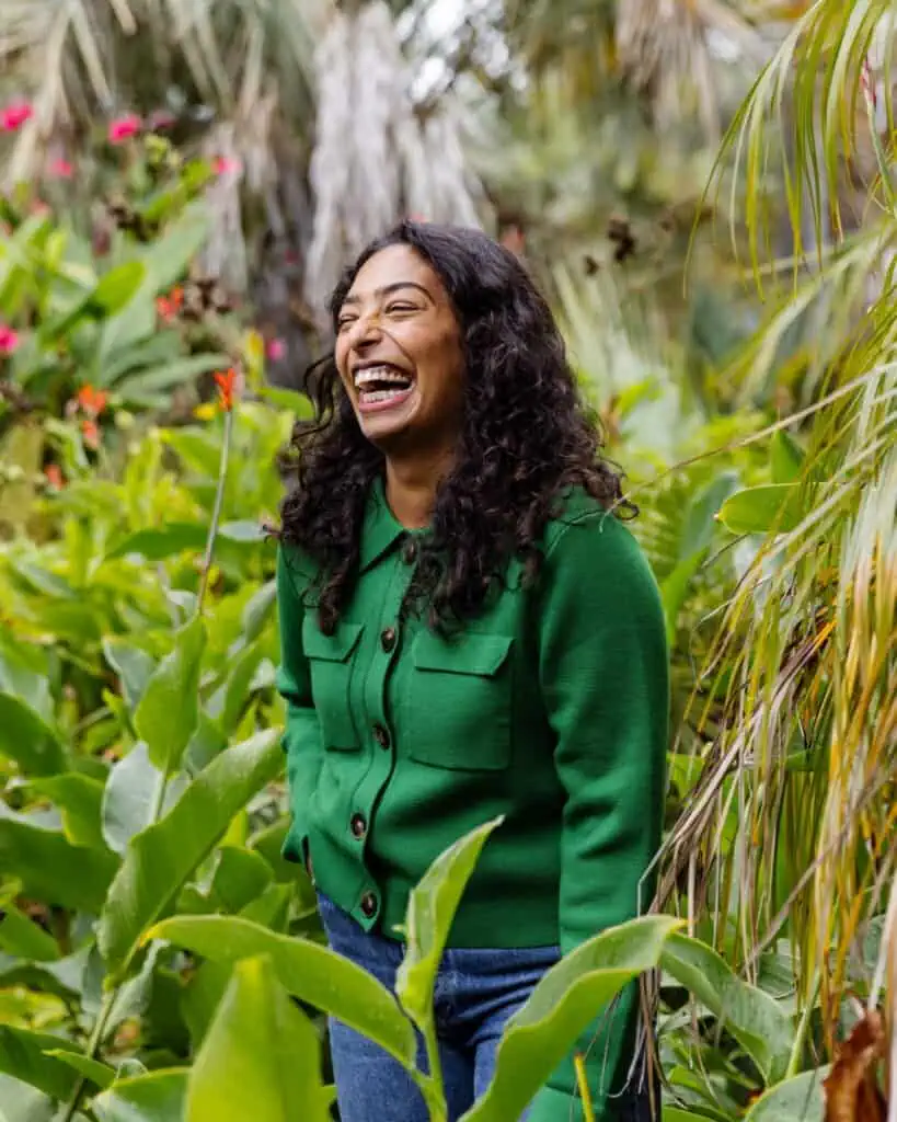 A woman with curly hair wearing a green jacket and blue jeans stands smiling and laughing in a lush, green garden with tropical plants and pink flowers.