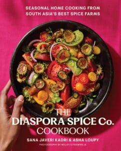A hand holds a black bowl filled with a colorful salad of tomatoes, cucumbers, onions, peppers, and herbs, set on a vibrant pink background. The cover text reads: The Diaspora Spice Co. Cookbook.