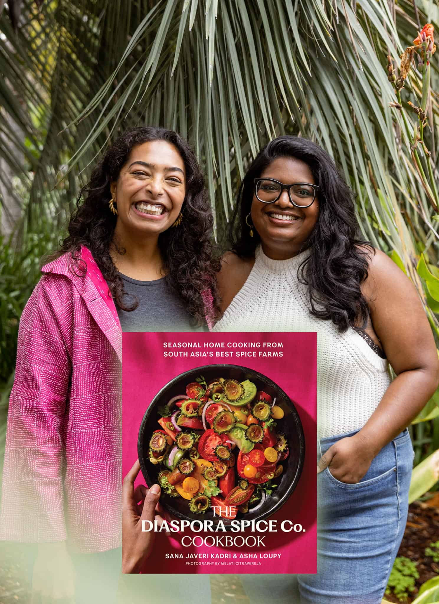 Two women stand smiling next to each other in front of green foliage. Beside them is the cover of The Diaspora Spice Co. Cookbook, featuring a colorful dish and text about South Asian home cooking.