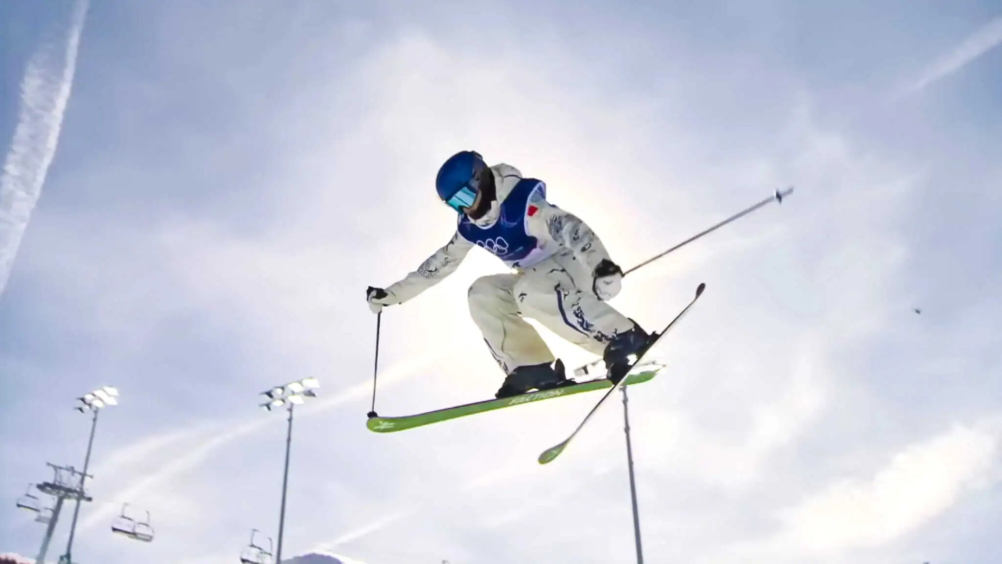 A skier in mid-air performs a jump against a bright sky, wearing a white suit, blue vest, helmet, and goggles, with ski poles outstretched and green skis angled beneath them.