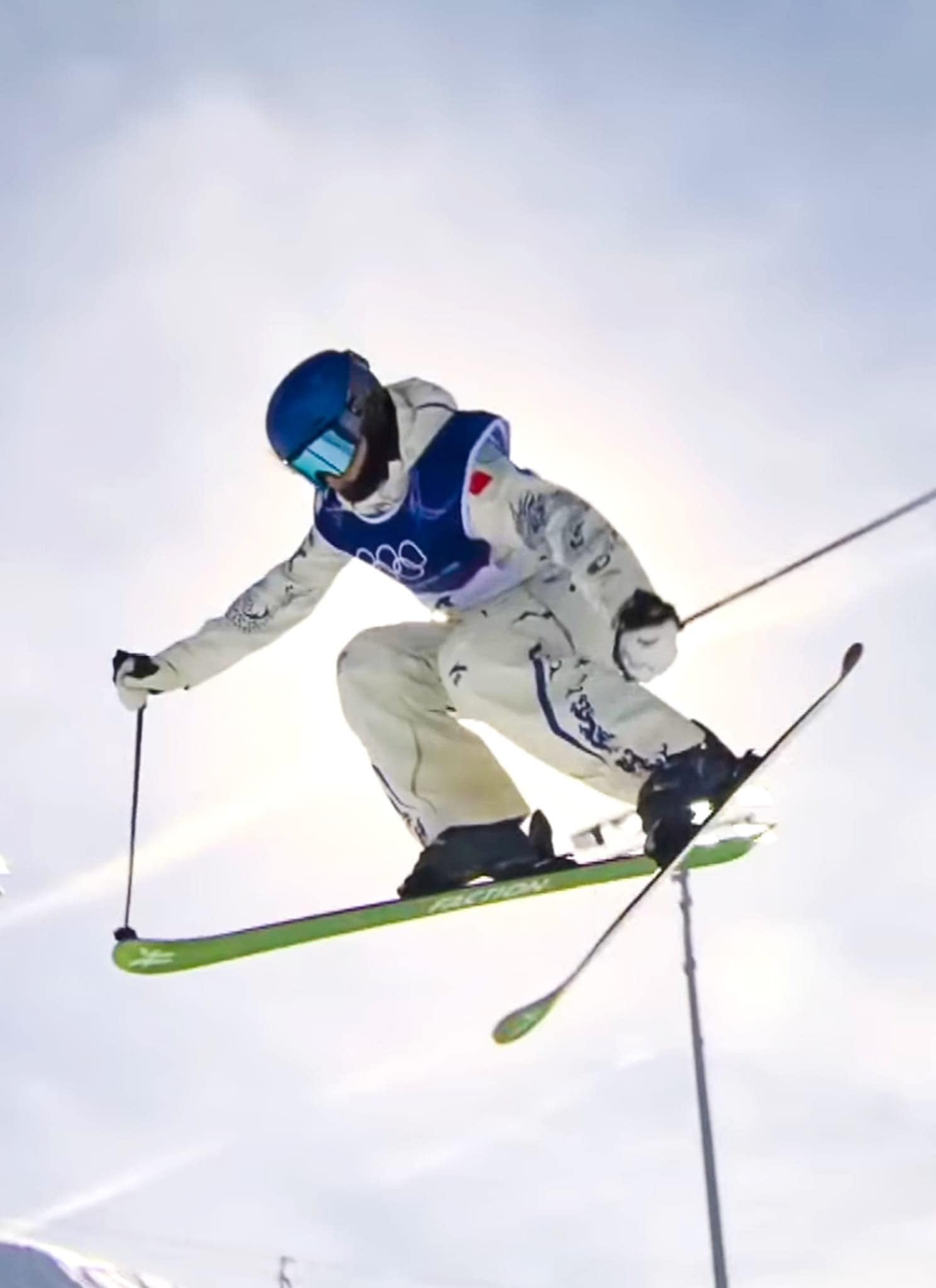 A skier in mid-air performs a jump against a bright sky, wearing a white suit, blue vest, helmet, and goggles, with ski poles outstretched and green skis angled beneath them.