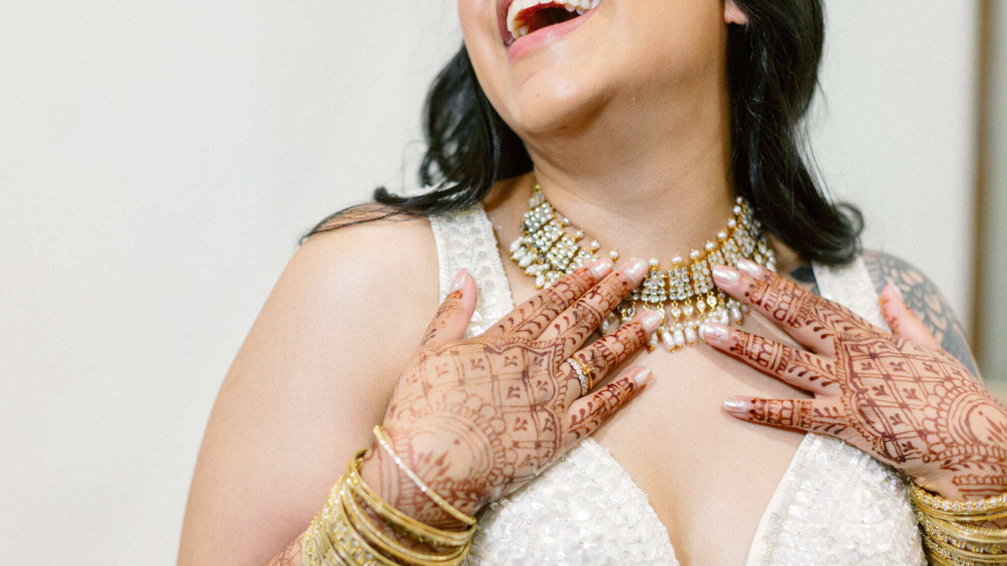 A woman wearing a jeweled necklace and gold bangles smiles, showing intricate henna designs on her hands. She is dressed in a white, embellished outfit.