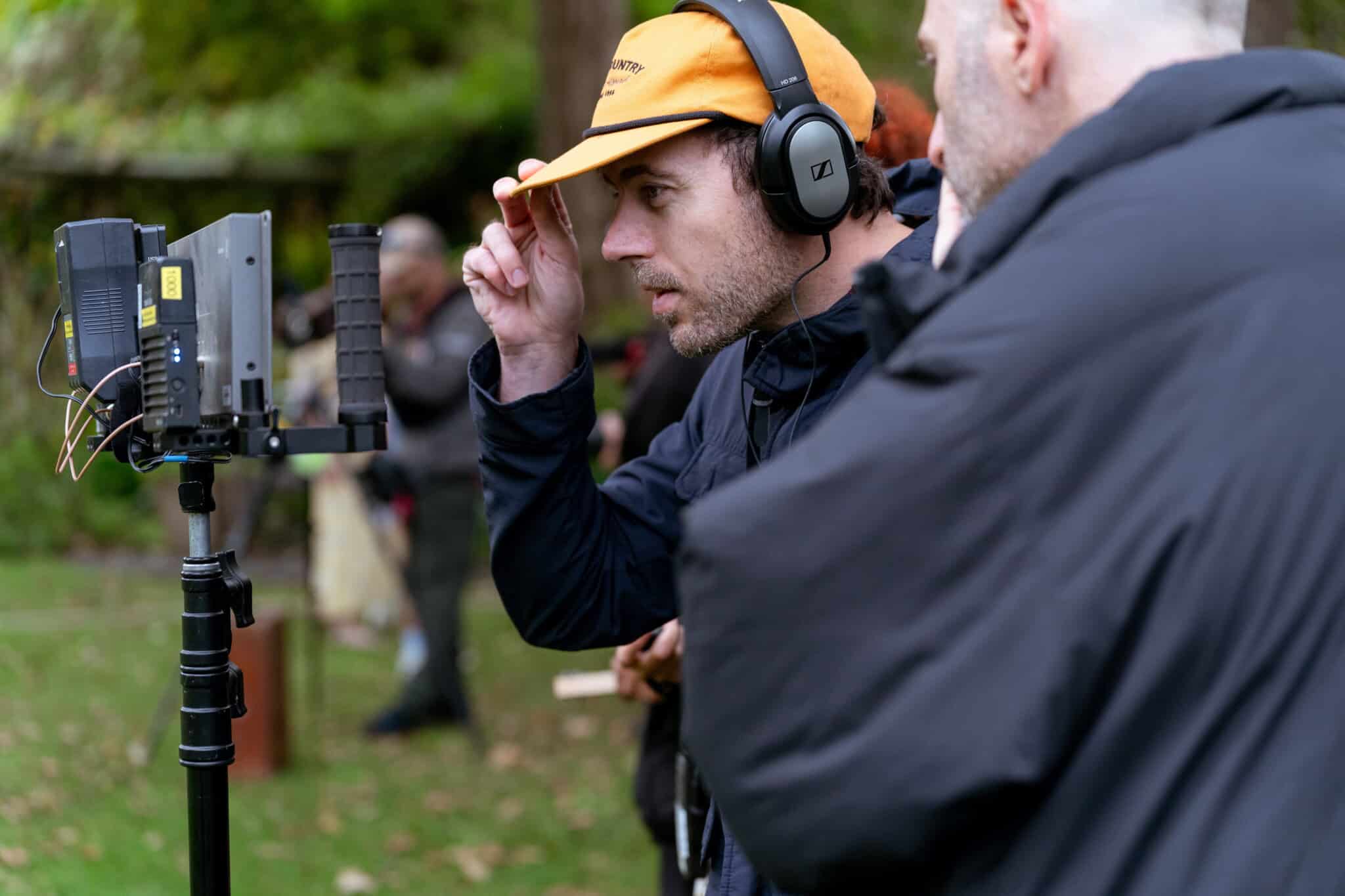 A man wearing headphones and an orange cap adjusts his hat while watching a monitor outdoors, with another person standing beside him. The scene appears to be part of a film or video production set.