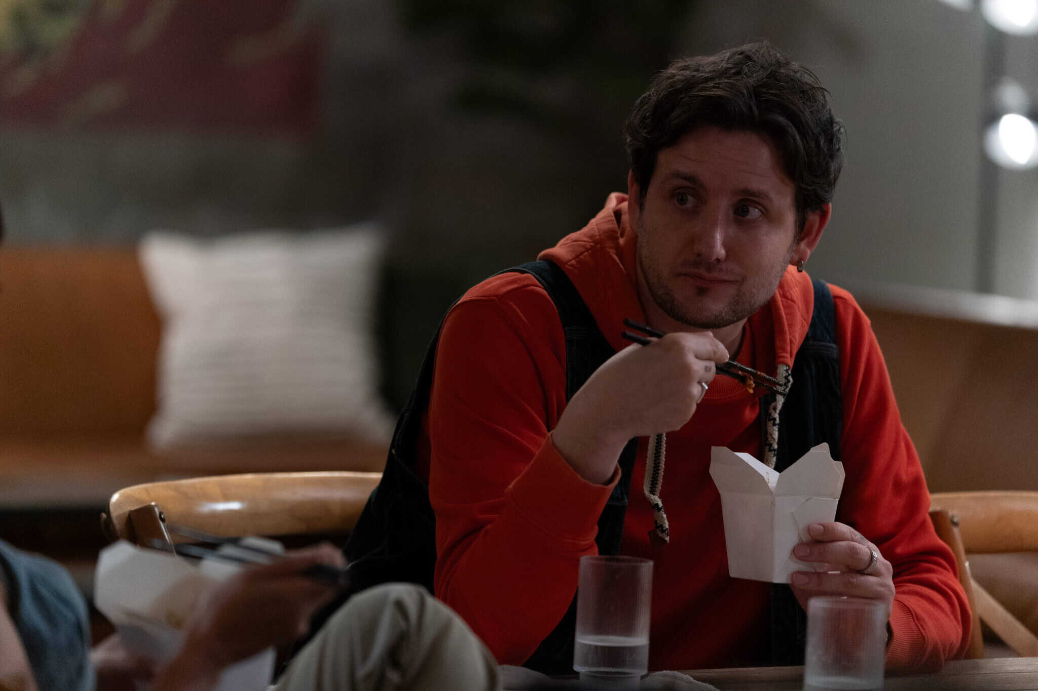 A man in a red hoodie uses chopsticks to eat from a white takeout box while sitting at a table. He is looking to the side and there are two glasses of water on the table.