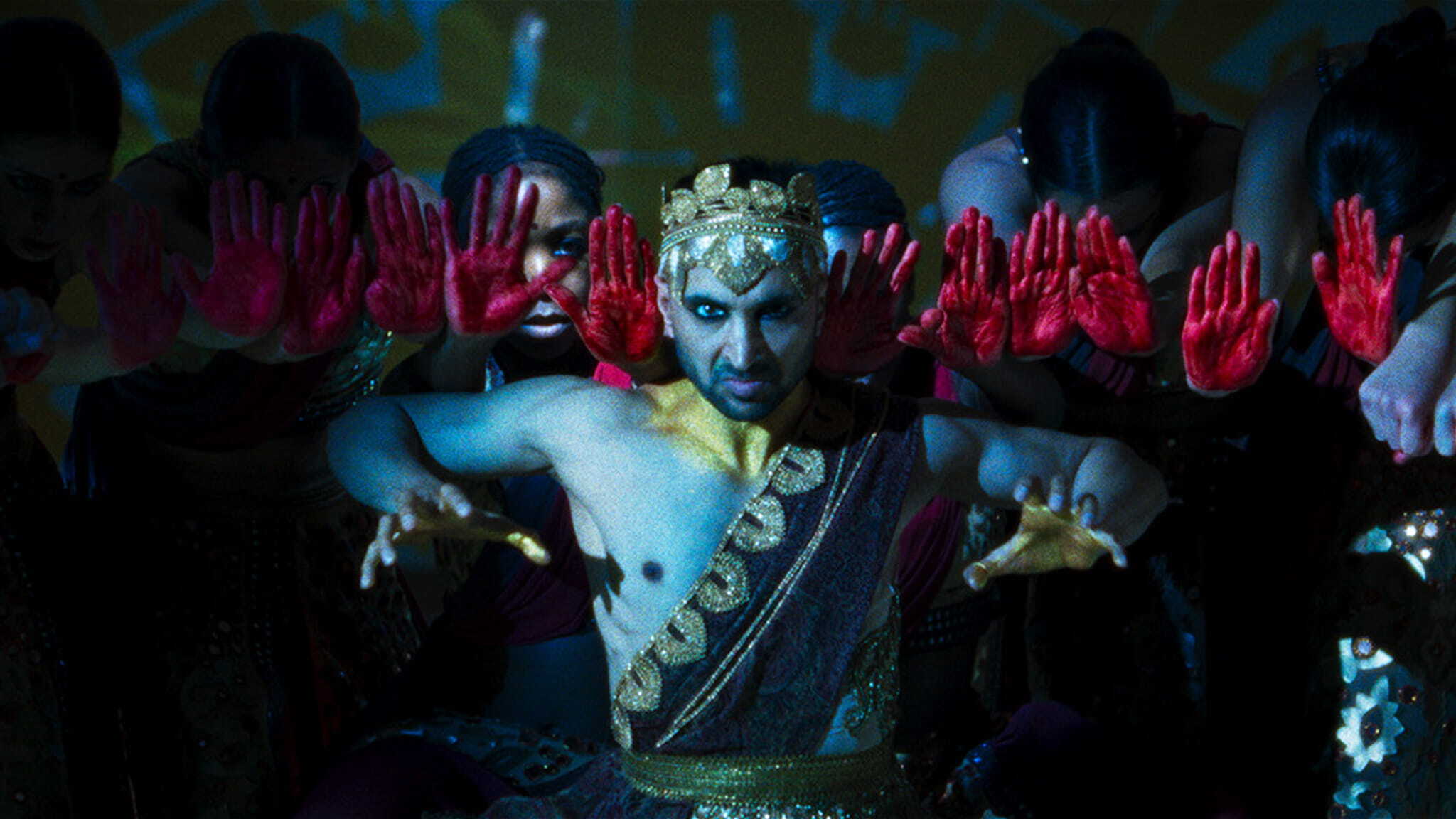 A performer in ornate golden attire and crown poses with outstretched hands, flanked by dancers holding up palms painted red, evoking a dramatic, synchronized stage performance with vivid lighting.
